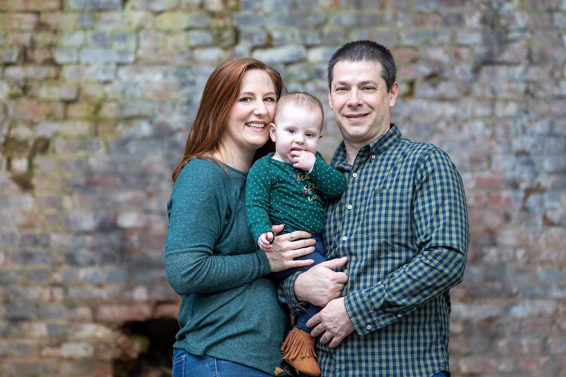 Portrait photo of couple all dressed in green holding their baby with a brick wall in the background