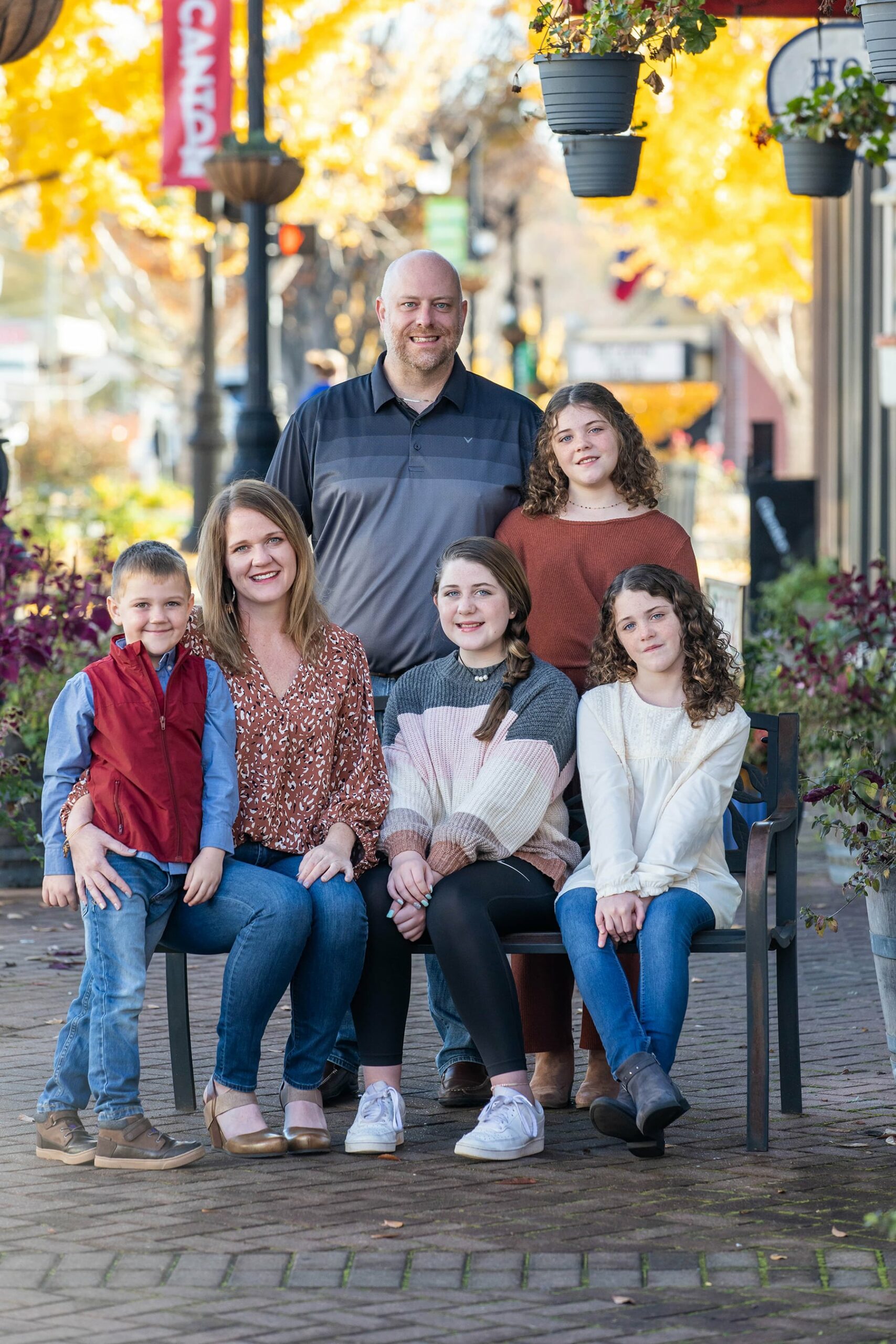 Family of six with the mother and her three kids sitting on a bench and others standing with picturesque downtown street in the background