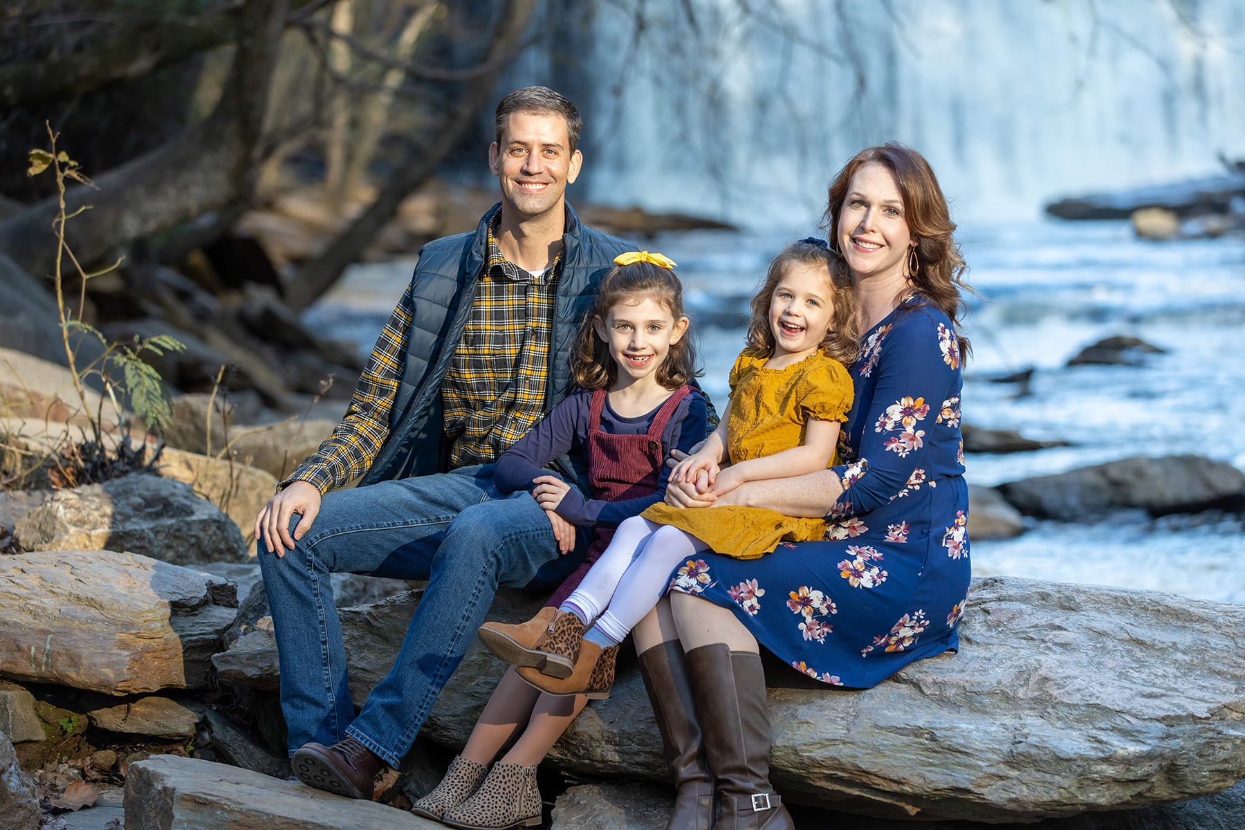 A family of four consisting of dad, mom and their two daughters sitting on a large stone with waterfall in the background
