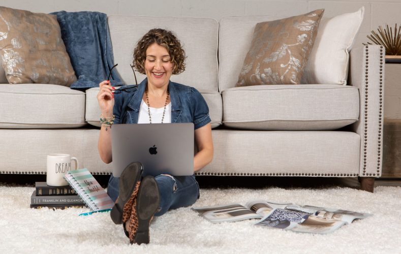 Photo of a woman smiling while looking at her laptop with books and magazines spread around